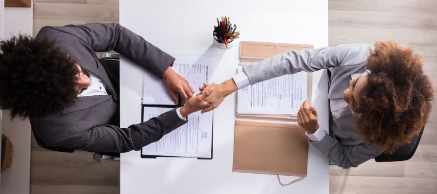Two business professionals from HR recruting consultancy shaking hands at a desk during a meeting.